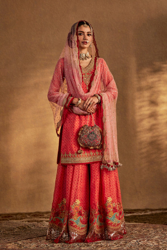 Model in a coral peach sharara set, vibrant embroidered skirt, matching top, traditional South Asian formal wear, elegant.