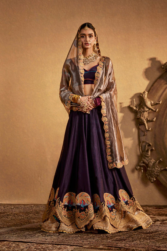 Model in an elegant deep purple lehenga with gold embroidery, traditional South Asian bridal wear, flowing skirt, ornate.