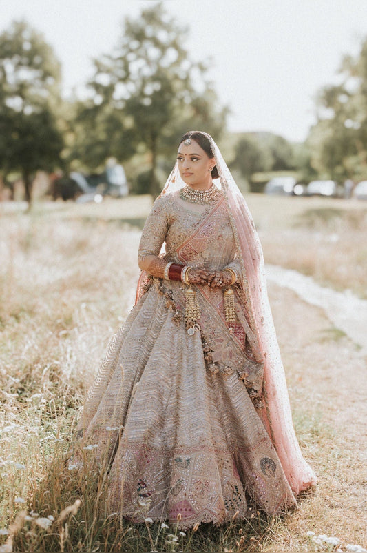 Indian bride in a muted gold and red bridal lehenga with traditional jewelry, standing in a stone corridor, RAH Weddings evening photography.