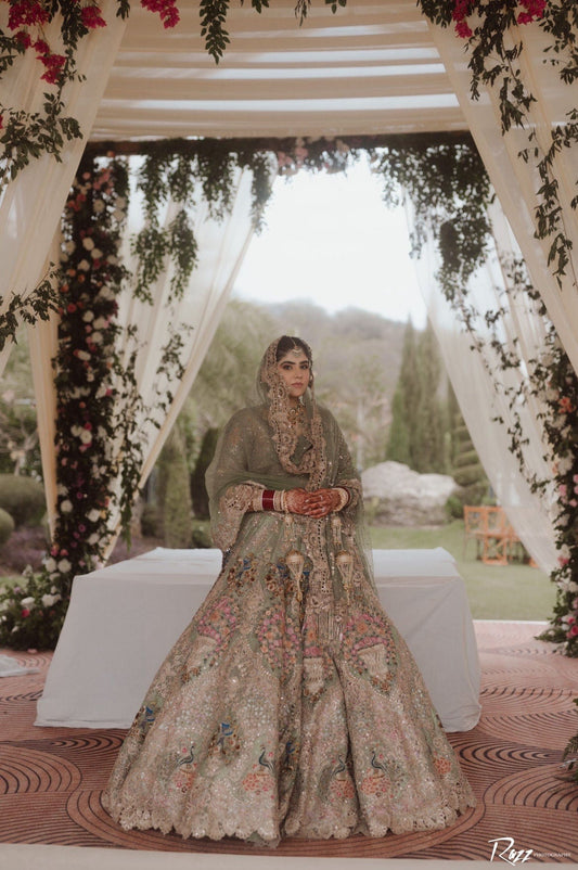 Elegant Indian bride in a heavily embroidered red and gold bridal lehenga, seated indoors with floral decor, RAH Weddings bridal portrait.