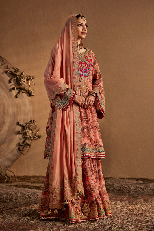 Model in an elegant blush peach sharara set with floral embroidery, traditional South Asian formal wear, flowing fabric.