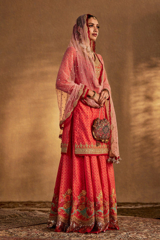 Model in a coral peach sharara set, vibrant embroidered skirt, matching top, traditional South Asian formal wear, elegant.